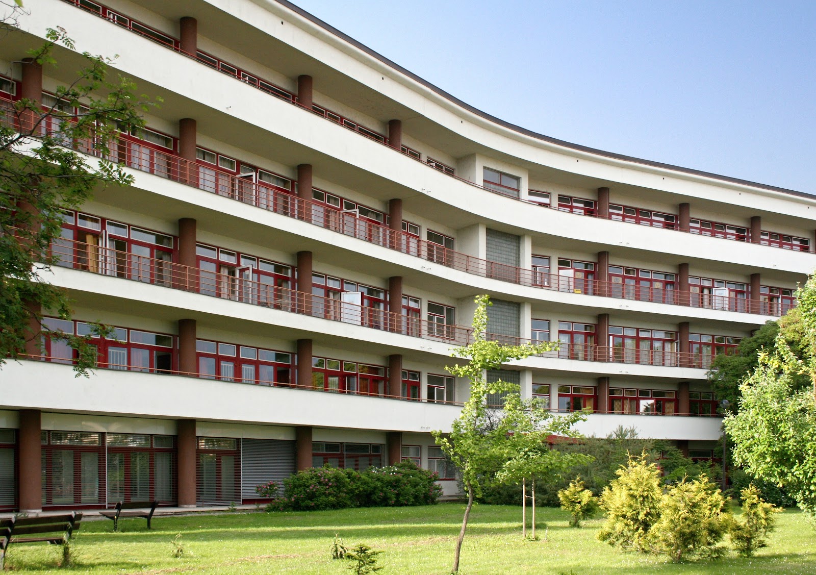 Zakřivená budova nemocnice s balkony a zelenou zahradou.